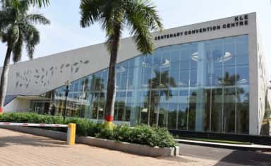 Glass-fronted KLE Centenary Convention Centre with its name signage and palm-lined forecourt, listed by JNMC Belagavi as campus infrastructure