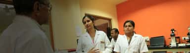 Postgraduate medical students in white coats discussing with a professor in a microscopy lab at KMC Mangalore