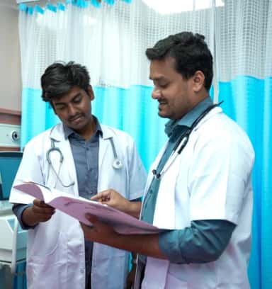 Two doctors in white coats reviewing a case sheet beside ward curtains at Takshashila Medical College Hospital