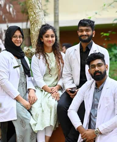 Four Yenepoya Medical College students in white coats posing under a tree on the Deralakatte campus, Mangalore