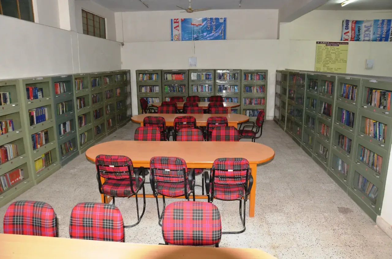 A study area in a library with green metal bookshelves lining the walls and red plaid chairs around wooden tables. — Arya College of Engineering & Research Cent