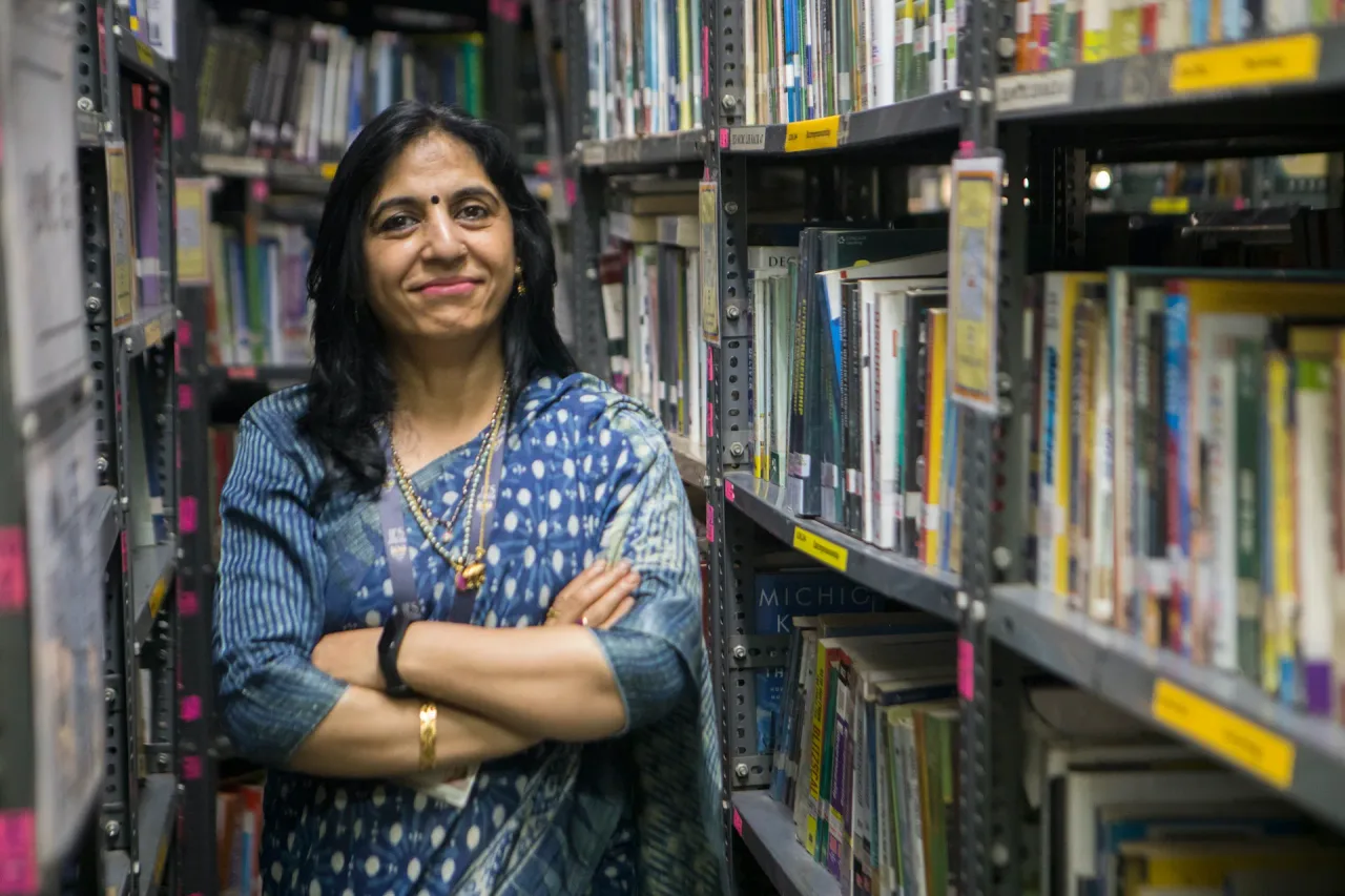 Smiling woman in blue patterned sari standing with folded arms between bookshelves in a library. — ies-s-management-college-and-research-centre-mumbai